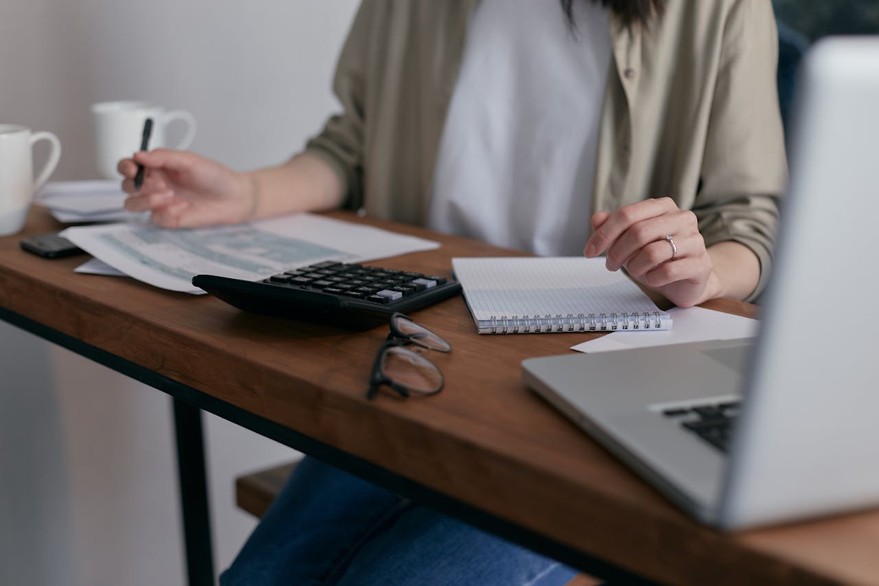 heros-img A woman manages finances at home, using a laptop and calculator on a wooden desk.